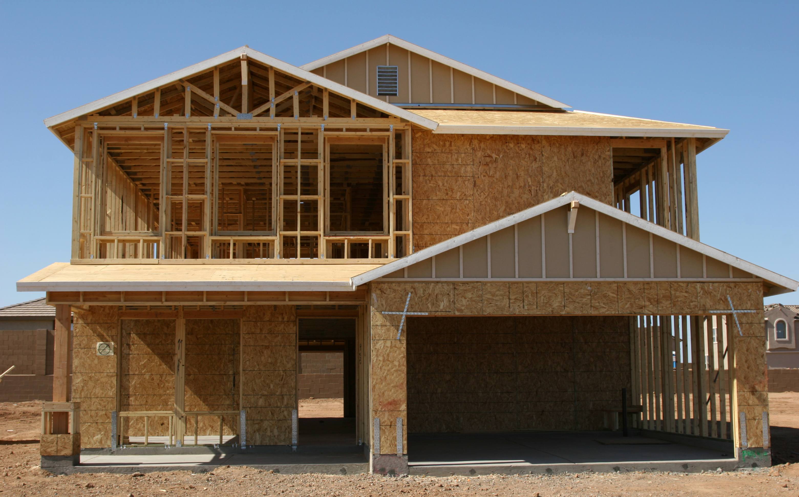 A two-story house under construction with a wooden frame, exposed plywood walls, and an unfinished roof against a clear blue sky.