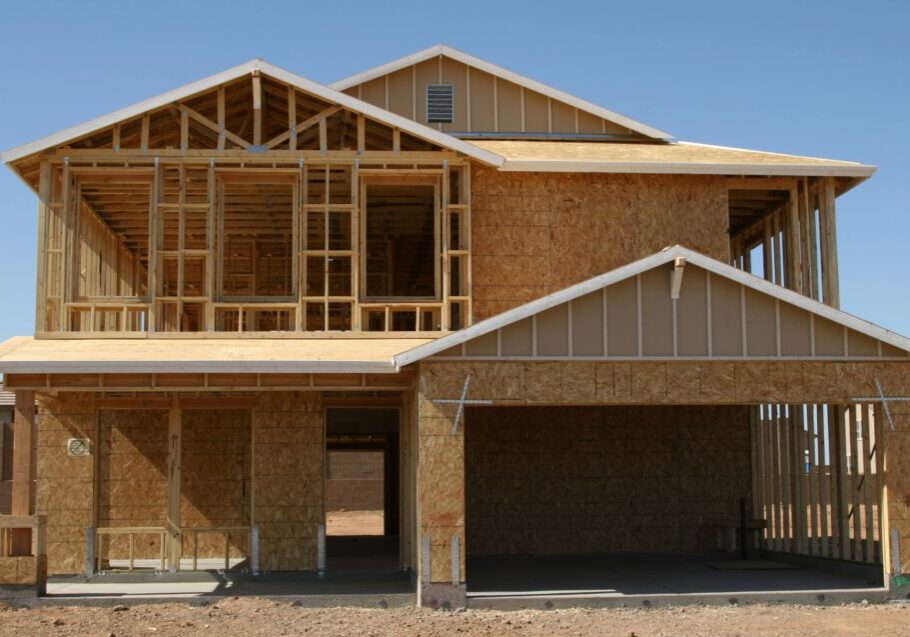 A two-story house under construction with a wooden frame, exposed plywood walls, and an unfinished roof against a clear blue sky.